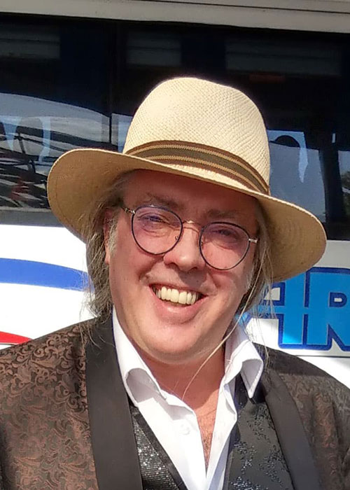 John Picton hat einen Panamahut, eine kastanienbraune Jacke und eine Brille auf